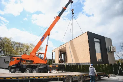 A crane lifts a manufactured house, with is rectangular and does not yet have a roof.