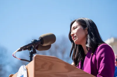 Mayor Wu speaks behind a wooden podium. She has black hair slightly past her shoulders and is wearing a magenta jacket. 