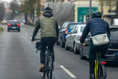 Two people on bikes in puffer jackets riding on street next to row of parked cars.
