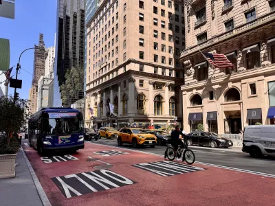 A bus lane with a bus driving on it and a person biking. Parallel, people drive cars down a busy street.