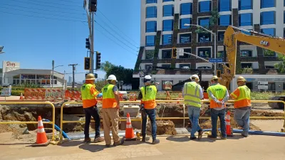 Department of Water and Power workers at the site of a water main rupture on Sunset Boulevard in West Hollywood. There are six men in construction vests looking at the rupture, with a crane in the background.
