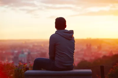 View from back of young man sitting on concrete bench overlooking blurred city at sunrise or sunset