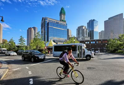 Person on bike wearing backpack with small bus and Boston buildings in background 