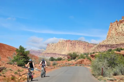 Two cyclists on road with red rocks of Capitol Reef National Park in background.