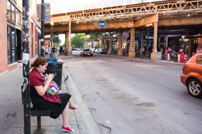 A woman sits on a bus stop bench and looks at her phone.