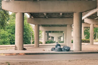 Man with grey hair sits next to black garbage bags in freeway underpass in Houston.