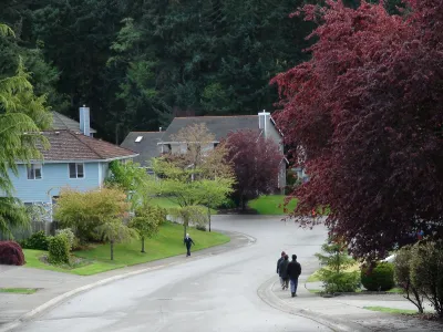 Three people walking or jogging on sidewalks in residential neighborhood with large homes and leafy trees.