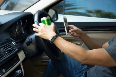 Close-up of man's arms in driver's seat with one hand holding green can on steering wheel and other hand holding cell phone.