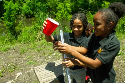 Two young Black girls outdoors using red plastic cup to pour water into tube for measurement or testing.