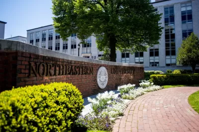 Image of the brick sign of Northeastern University's sign and logo with a small garden and brick pathway in front of it and white buildings in the background