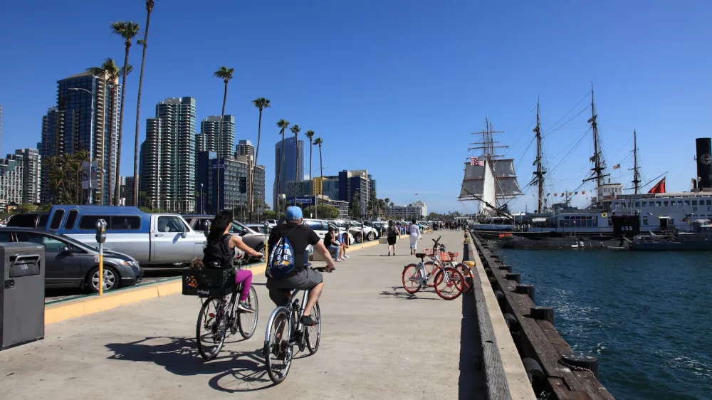 People biking along beach path with moored ship in San Diego, California.