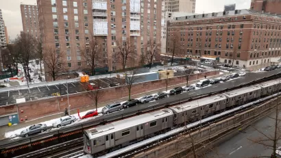 Silver New York City subway train on above ground track with red brick multistory buildings in background.