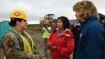 Three people, one in fatigues and hard hat, standing in front of bulldozer discussing relocation plans in Newtok, Alaska.