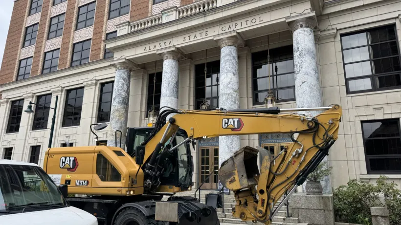 A CAT excavator drives past a brick building with marble columns.
