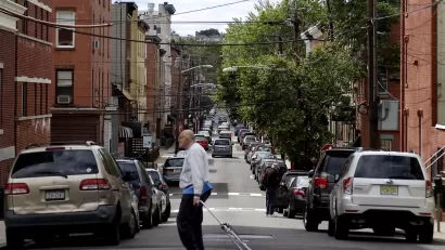 An old man crosses a one-way street in the foreground, with both sides of the streets lined with cars facing away from the camera and brick buildings.