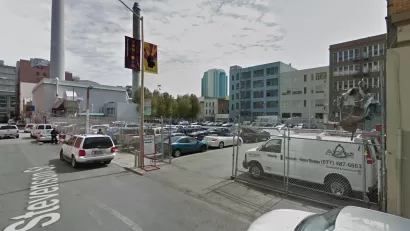 A surface parking lot, full of cars, is located between tall buildings and urban density.