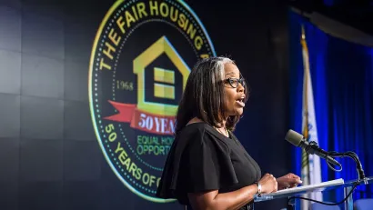 Woman speaks at lectern in front of banner celebrating the 50th anniversary of the Fair Housing Act in 2018.