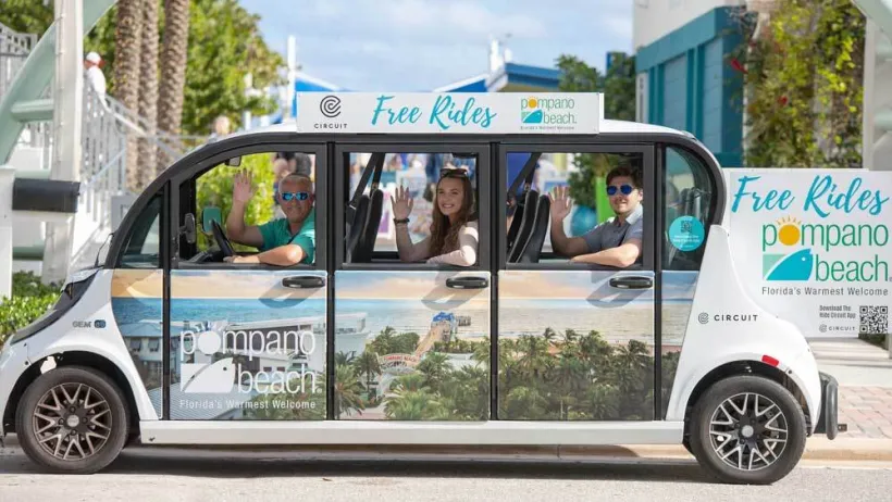 Small white microtransit shuttle with wrap depicting beach town in Pompano Beach, Florida with happy people waving from inside.