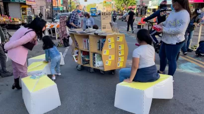 Children sit on temporary street furniture next to book cart at pop-up reading event at open streets event in Sunset Park in New York City.