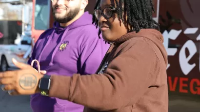 Black woman in brown sweatershirt and Black man in purple sweatshirt stand in front of food truck.