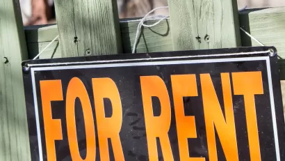 Close-up of orange and black For Rent sign on green wood fence.