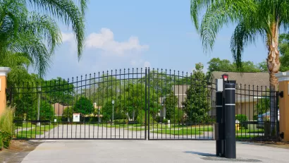 Closed black wrought iron gate in front of gated residential community with large palm trees along sides of street.