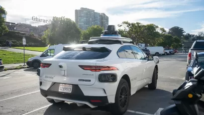 White Waymo autonomous vehicle in parking area in San Francisco, California with Ghirardelli vintage sign in background.
