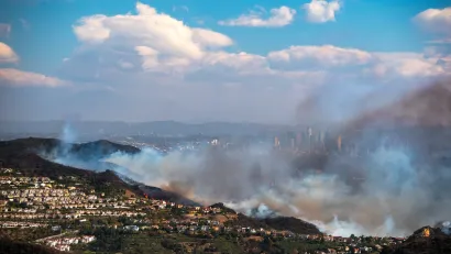 Heavy smoke from Palisades Fire burning in hillsides with homes with downtown Los Angeles visible in background.