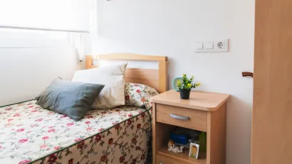 Small, brightly lit bedroom with flowery sheets on single bed in supportive housing or nursing home.