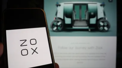 Close-up of smartphone with Zoox logo and screen with blurred image of Zoox autonomous vehicle in background.