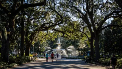 Paved path with artesian fountain at end and tall trees arching over walkway with joggers and walkers in Savannah, Georgia.
