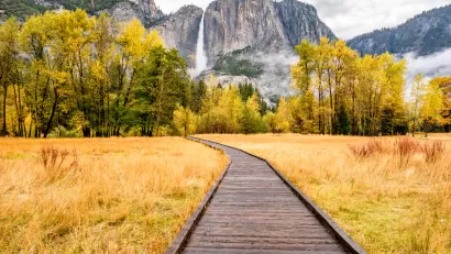Wooden boardwalk through yellowed grass field with tall granite cliffs and waterfall in background in Yosemite National Park, California.