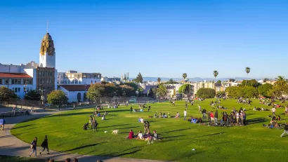 View of people sitting in lightly sloped green lawn with Spanish colonial-style building and cityscape in background in San Francisco, California.