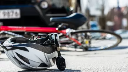 White bike helmet lies upturned on a road with red bicylce underneath the front of a far in the background.