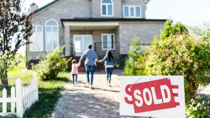 'For sale' sign with 'Sold' update in front of large single-family home with family of three walking toward house.