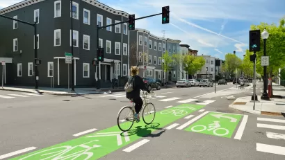 Cyclist on bike in green painted bike lane at intersection with three-story buildings in background.