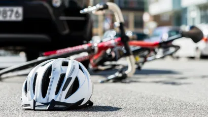 Close-up on white bike helmet lying on pavement with blurred red bike on its side in background abd black car visible behind it.