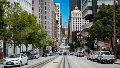 View from middle of street with streetcar rails and high-rise buildings on either side in San Francisco, Bay Bridge visible in background