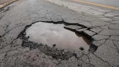 CLose-up of pothole in asphalt road filled with water reflecting grey cloudy sky.