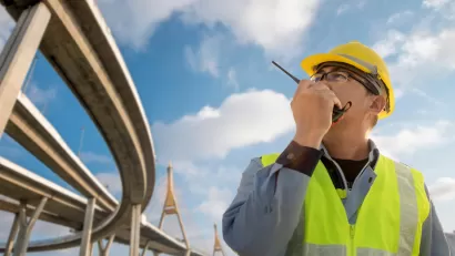 Civil engineer in yellow hard hat and safety vest talks on radio while looking up at freeway ramps.