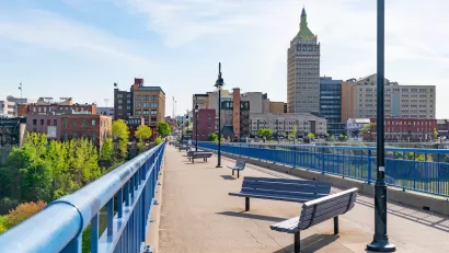 Pedestrian bridge with benches in Rochester, New York with brick buildings in background.