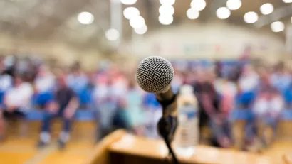Close-up of microphone on speaker's dais with blurred crowd sitting in bright high school gym in background.