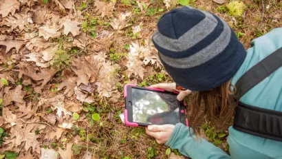 Woman wearing beanie leaning down over grass covered with fall leaves using tablet to identify plants.