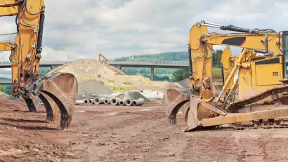 Three yellow excavators at a road construction site with elevated roadway in background.