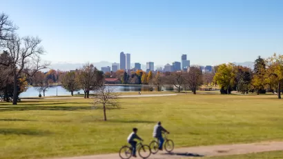 Two people on bikes on trail on green lawn with Denver, Colorado skyline in background.