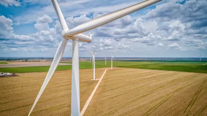 A line of white wind turbines surrounded by wheat and soybean fields with a cloudy blue sky in the background.