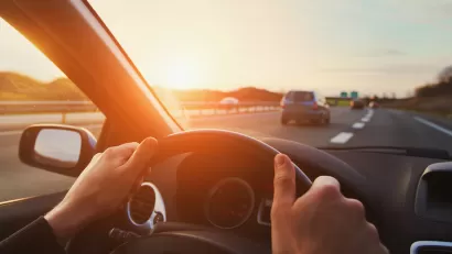 Cose-up of person's hands on steering wheel driving down highway with sunset sky.