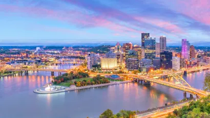 View of downtown Pittsburgh, PA with river and bridge in foreground at dusk.