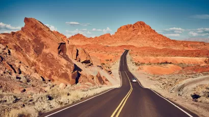 Two-lane highway with solid yellow center line and one car driving away against backdrop of red rock formations in Nevada.