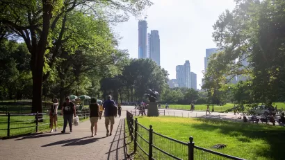 People walking on paved path in green city park with trees and tall city skyscrapers in background.